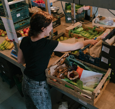 A women is in front of boxes of fresh fruit and veg, she's reaching over to pack the boxes. Her back is to the camera.