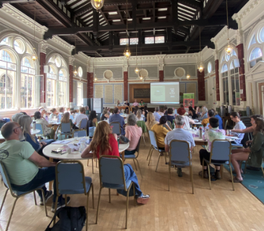 A group of traders sit in a big hall looking at a presentation. they're sitting around round tables